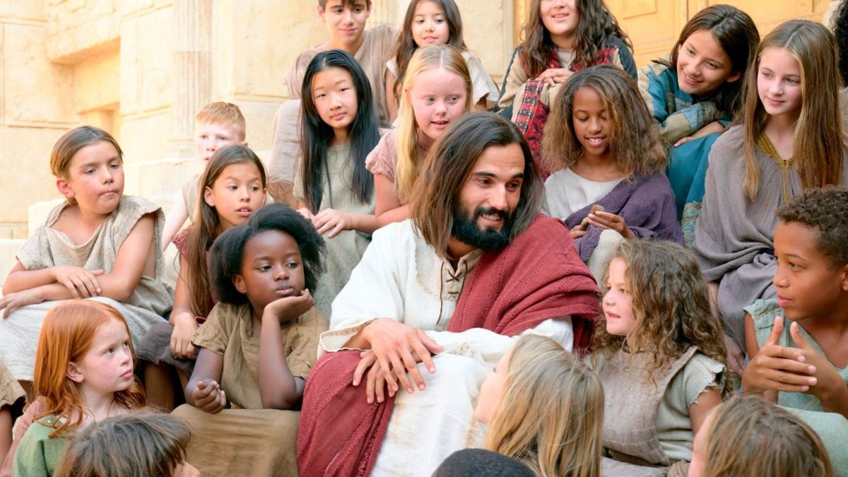 Jesus Christ seated on stone steps, smiling, surrounded by a crowd of children