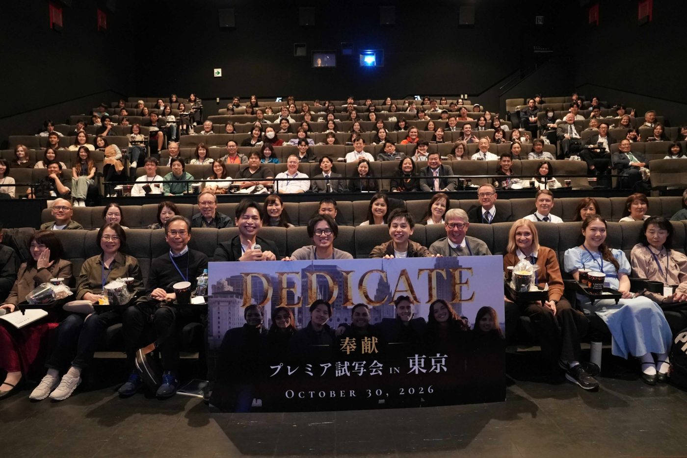 A full theater audience in Tokyo behind a banner for the Dedicate premiere