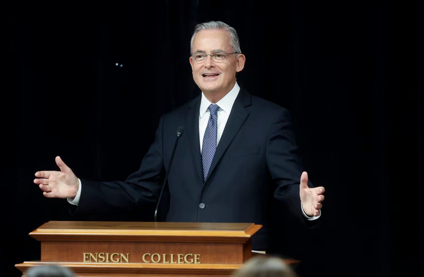 Elder Patrick Kearon smiling with arms open behind the Ensign College pulpit