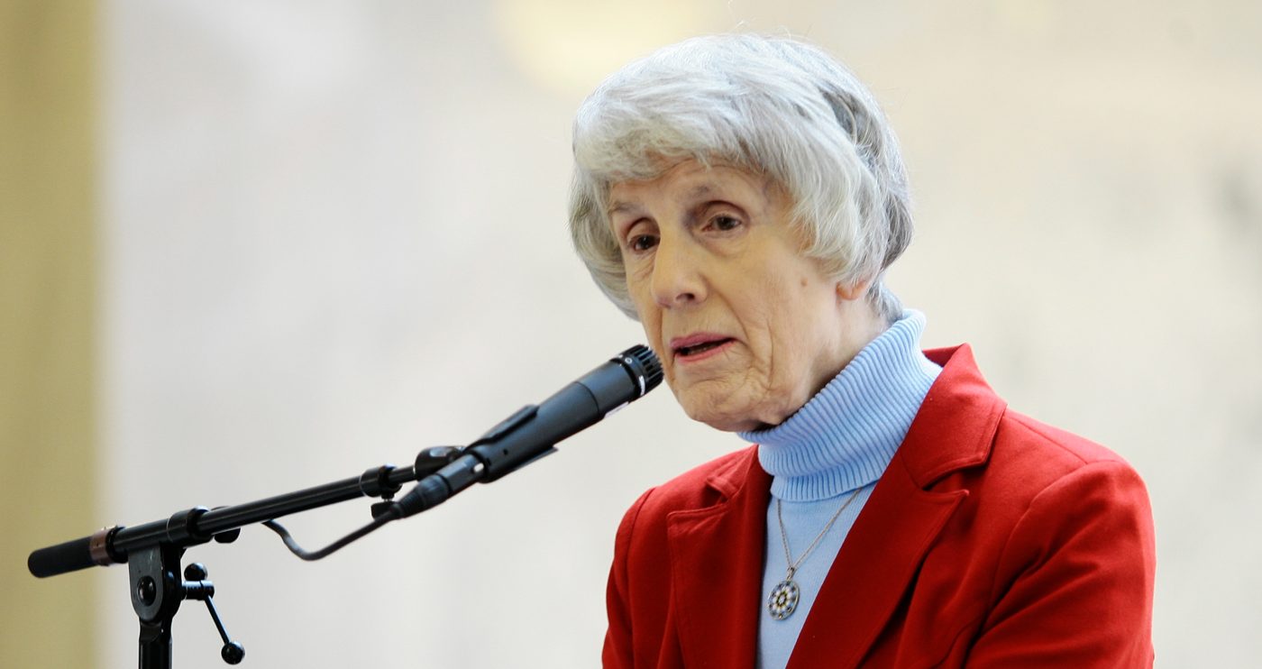 Pamela J. Atkinson speaking at a microphone in a red jacket