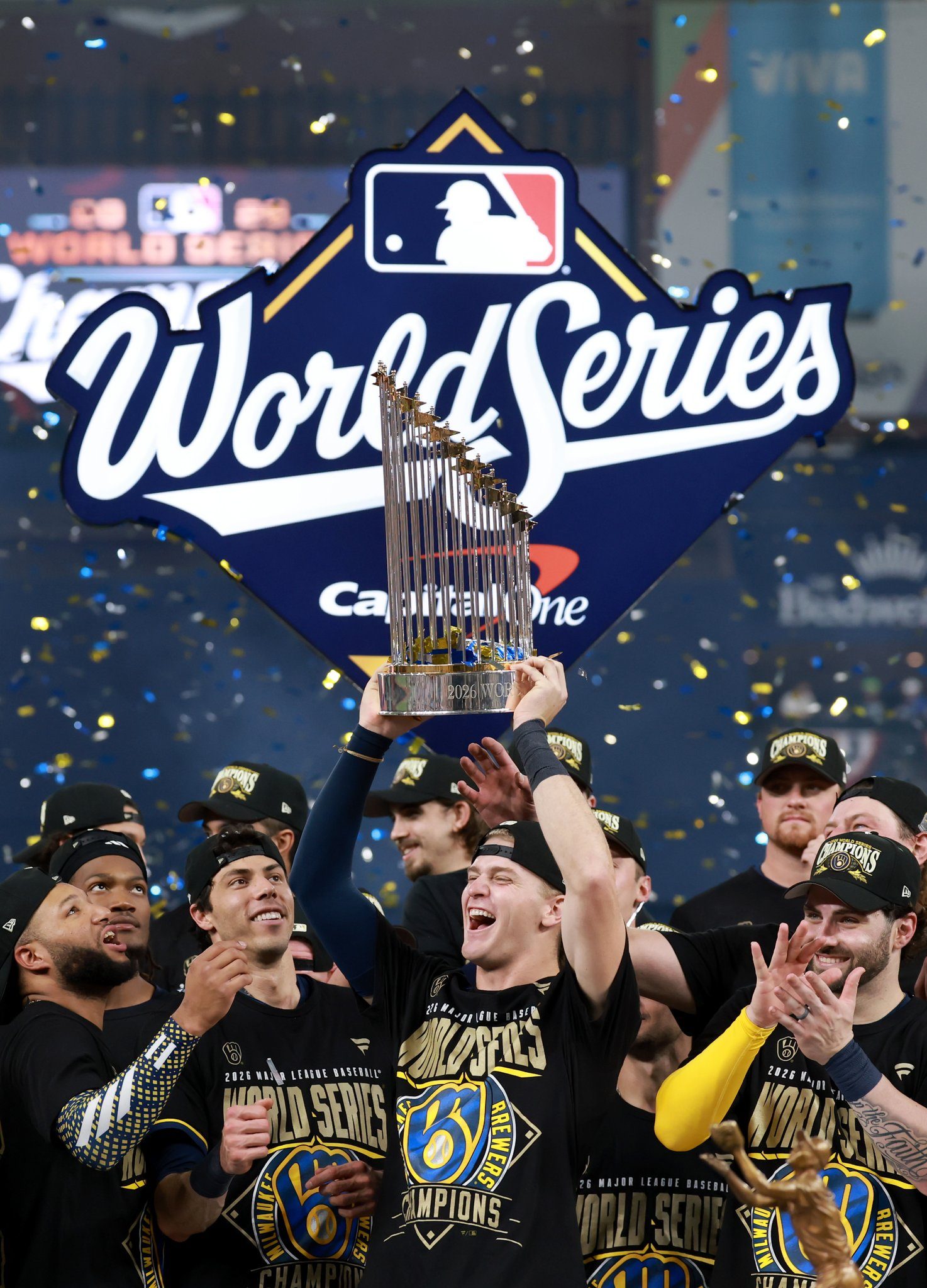 Cooper Pratt, in a World Series champions shirt and cap, cheering beneath the raised World Series trophy as confetti falls and teammates celebrate around him
